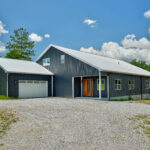 Front exterior of the barndominium-style home with dark metal siding, standing seam roof, wood and glass front door, attached garage, and gravel driveway