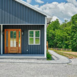 Exterior view of the home's covered front entry with dark metal siding, a wood and glass door, and a curved paver walkway leading to the gravel drive