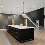 Kitchen with black island, white quartz countertop, gold pendant lighting, and a cable-railing staircase visible in the open living area beyond
