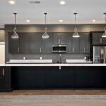 Kitchen island with four black and brass pendant lights facing a wall of black cabinetry, built-in microwave, and stainless refrigerator
