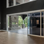 View through the open glider door to the covered back porch, showing a stone-faced see-through gas fireplace with wooded hillside beyond
