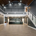 Two-story great room with vaulted tongue-and-groove ceiling, cable-railing staircase and upper catwalk, and a dark cabinetry kitchen with pendant lighting in the background