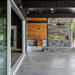 Covered back porch with a two-sided stone fireplace column, tongue-and-groove wood accent wall, dark metal ceiling, and a large glass sliding door reflecting the wooded yard