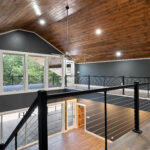 Upstairs catwalk with black cable railing and tongue-and-groove vaulted ceiling overlooking the wood-floored level below, with a wall of windows and a door to an upper deck