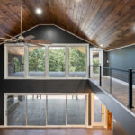 Vaulted tongue-and-groove ceiling with a wood ceiling fan above a catwalk with cable railing, multi-pane windows overlooking a deck, and a glass sliding door on the floor below