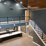 Elevated view from the catwalk down onto a kitchen with dark cabinetry, stainless appliances, and a light quartz island, with a cable-railing staircase and vaulted ceiling alongside