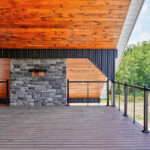 Covered upper deck with composite decking, black cable railing, tongue-and-groove wood ceiling, dark metal and wood siding, and a stone-faced column, overlooking a fenced pasture and trees