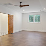 Empty bedroom with dark wood flooring, a vaulted ceiling with wood ceiling fan, a window looking out to trees, and open double wood plank doors leading to a hallway