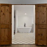 Arched wood double doors open into a bathroom revealing a freestanding white tub, matte black floor-mount faucet, and geometric patterned tile floor