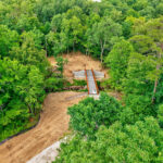 High aerial of the bridge centered in the wooded ravine, with the cleared approach, the curving new trail, and black silt fencing along the graded edge.