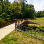 Wide aerial view of a wooded creek corridor with a bridge visible in the tree canopy, businesses and a residential area in the background