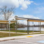 Angled view of both bocce ball court shelters with standing seam roofs, accessible parking signage, and the lake beyond