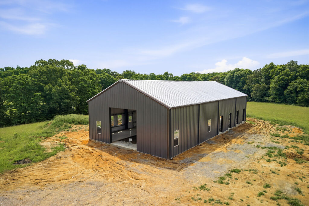 Aerial corner view of the charcoal steel barn with living quarters, showing the gable garage opening and the freshly graded clay pad around the slab.