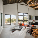 Open interior of the guest home and event space with a brown leather Chesterfield sofa, white swivel chairs, twin candle chandeliers, a dark stained vaulted plank ceiling, and a glass roll-up garage door opening to the patio.