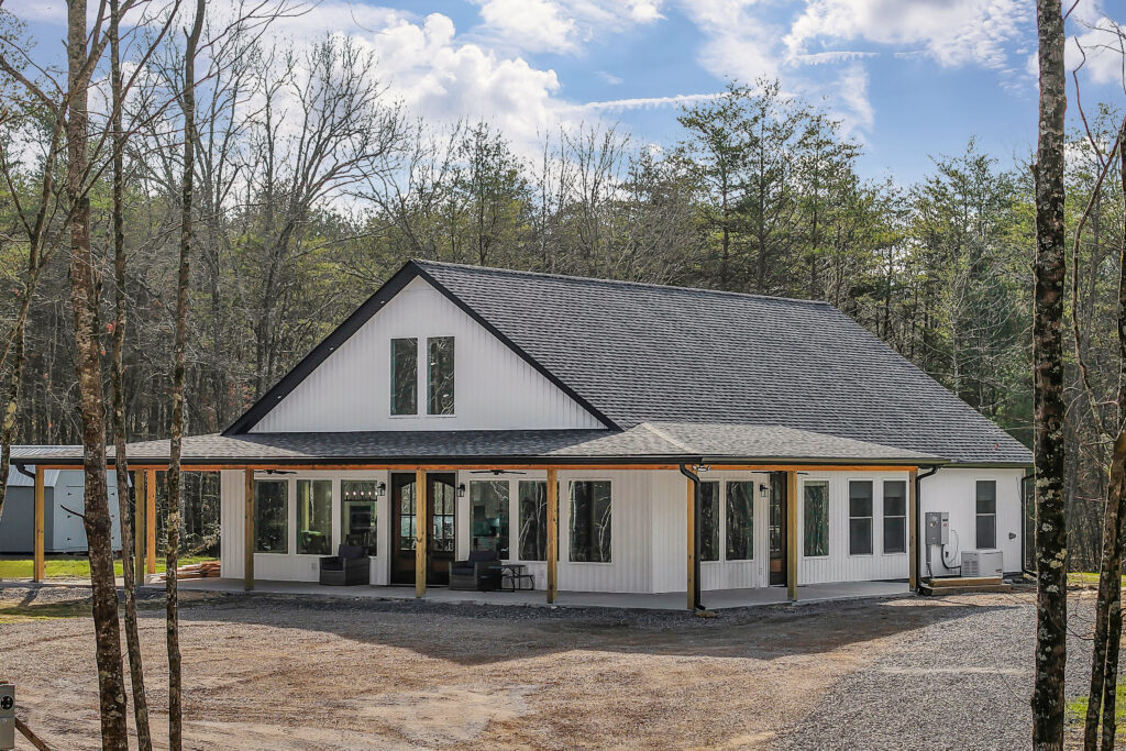 Aerial view of The Ridgeview farmhouse home with wraparound porch and board-and-batten siding on wooded land in Crossville, TN