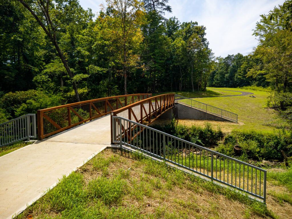 Wide aerial view of a wooded creek corridor with a bridge visible in the tree canopy, businesses and a residential area in the background