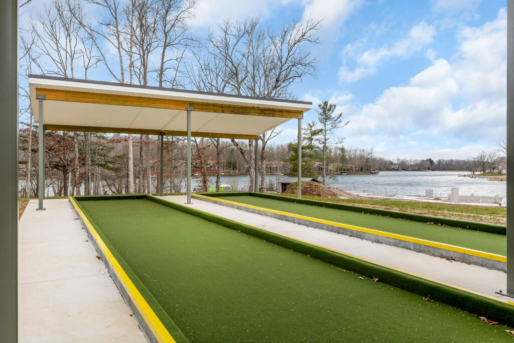 View down the bocce ball court lanes toward the lake, with sitework visible along the shoreline