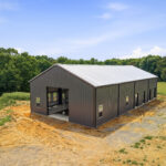 Aerial corner view of the charcoal steel barn with living quarters, showing the gable garage opening and the freshly graded clay pad around the slab.