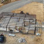 Aerial view of a steel building frame under construction, with exposed red steel columns and beams on a concrete slab, workers in safety vests, a scissor lift, and a further-along gray steel building beside it.