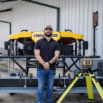 An East Tennessee Contractors team member stands with arms crossed in front of the company's new yellow rotary screed machine, with a laser level set up beside it.